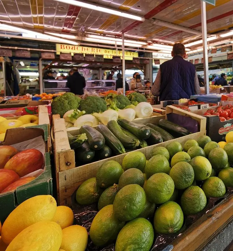 Caisses d'avocats, courgettes, brocolis et mangues sur un étal de marché couvert animé avec des clients en arrière-plan