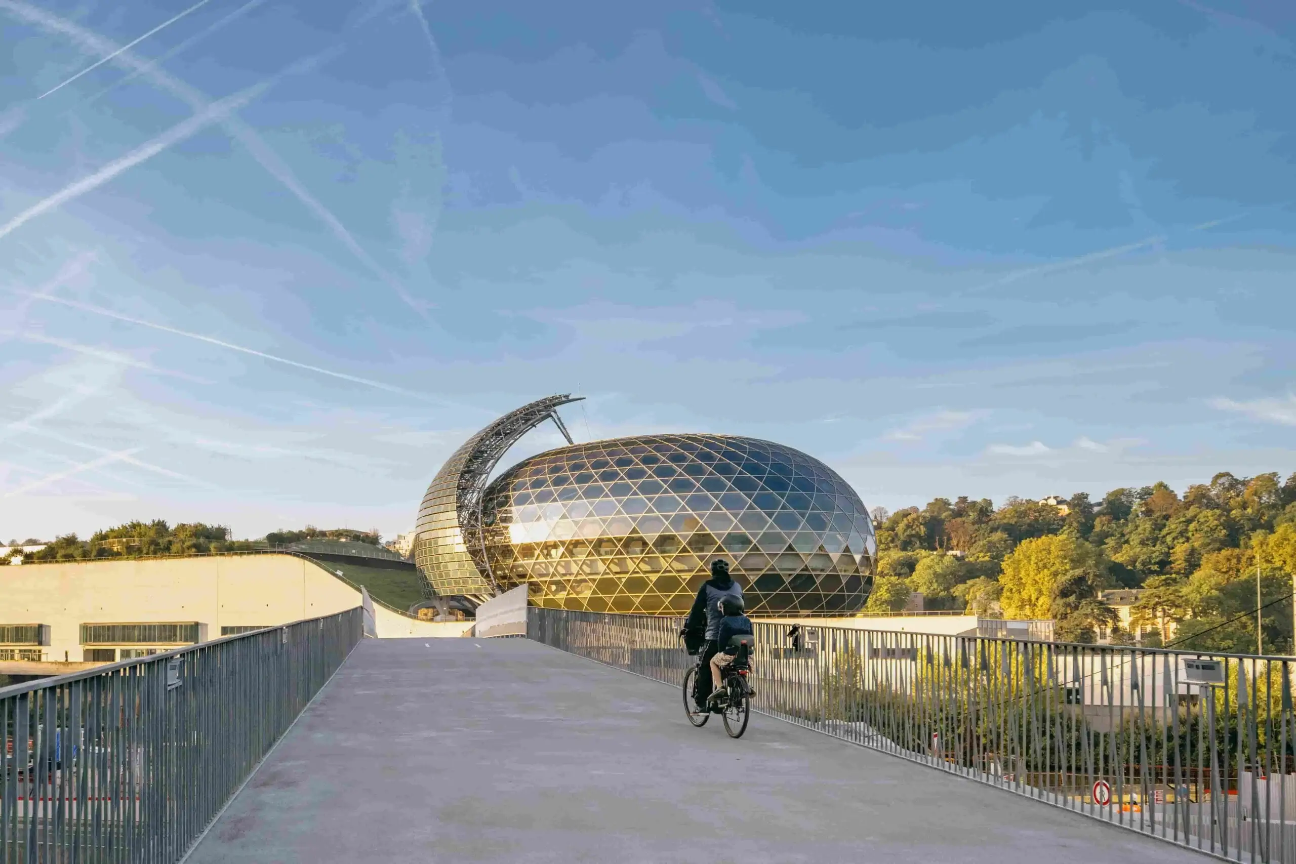 Cycliste sur une passerelle face à la Seine Musicale, bâtiment ovoïde à façade en verre et structure métallique dorée, entouré de verdure